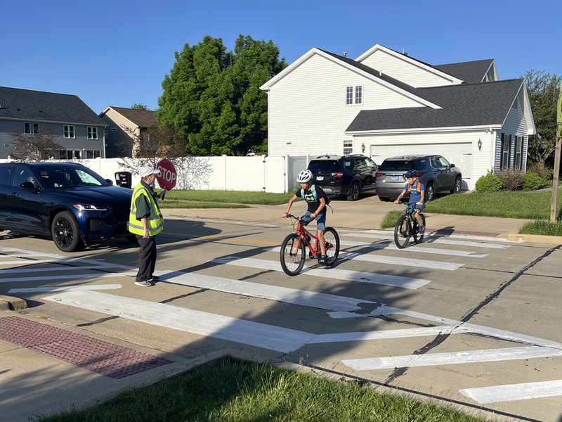 A crossing guard stops traffic while two children cross the street on their bicycles.